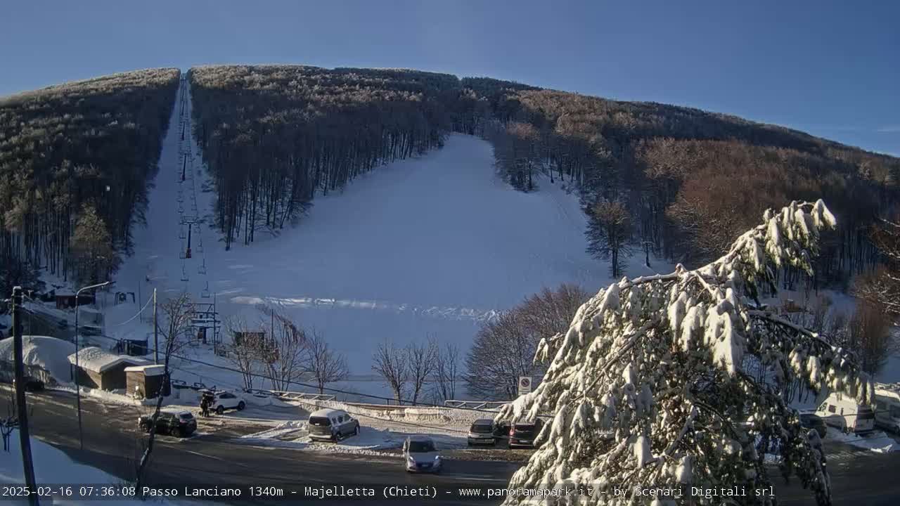 A snow-covered ski slope with a chairlift ascends a hill on a sunny day, with several cars parked in a lot below.
