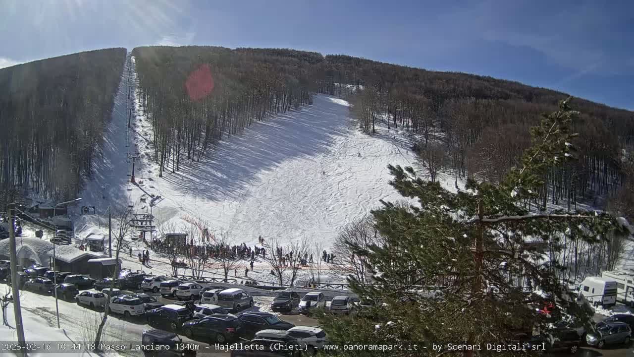 A snow-covered ski slope nestled in a wooded mountainside on a sunny day, with a parking lot full of cars at the base.