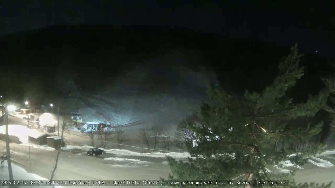 A nighttime view of a snow-covered parking lot and ski slope with a few cars and buildings visible under dark, clear skies.