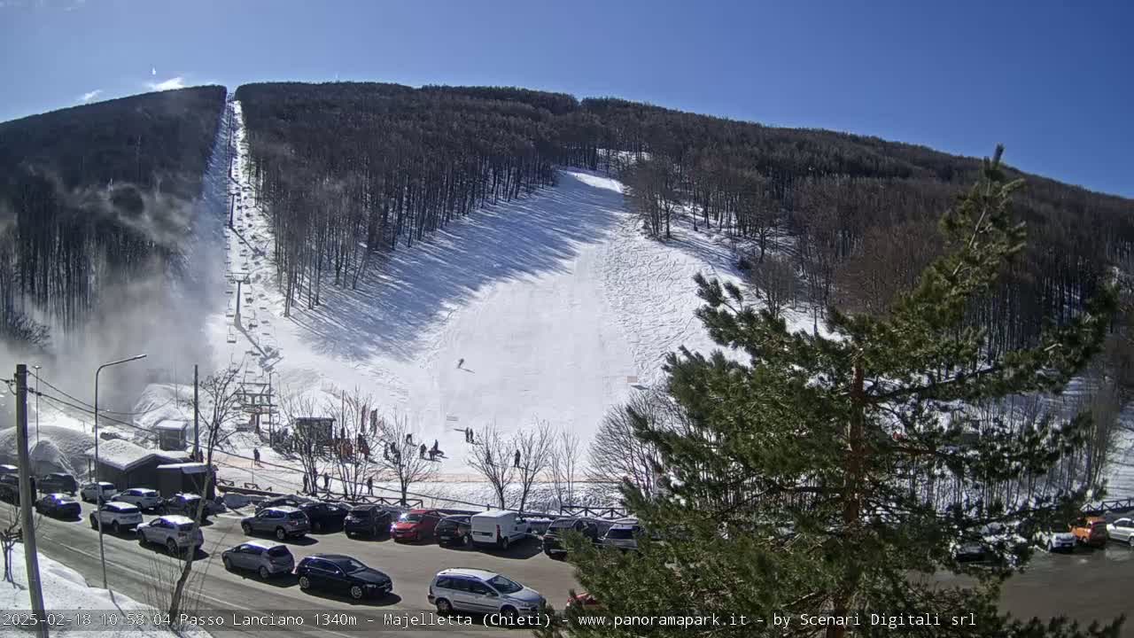 A snow-covered ski slope with a chairlift and several skiers, under a sunny sky, is viewed from a parking lot with numerous cars.