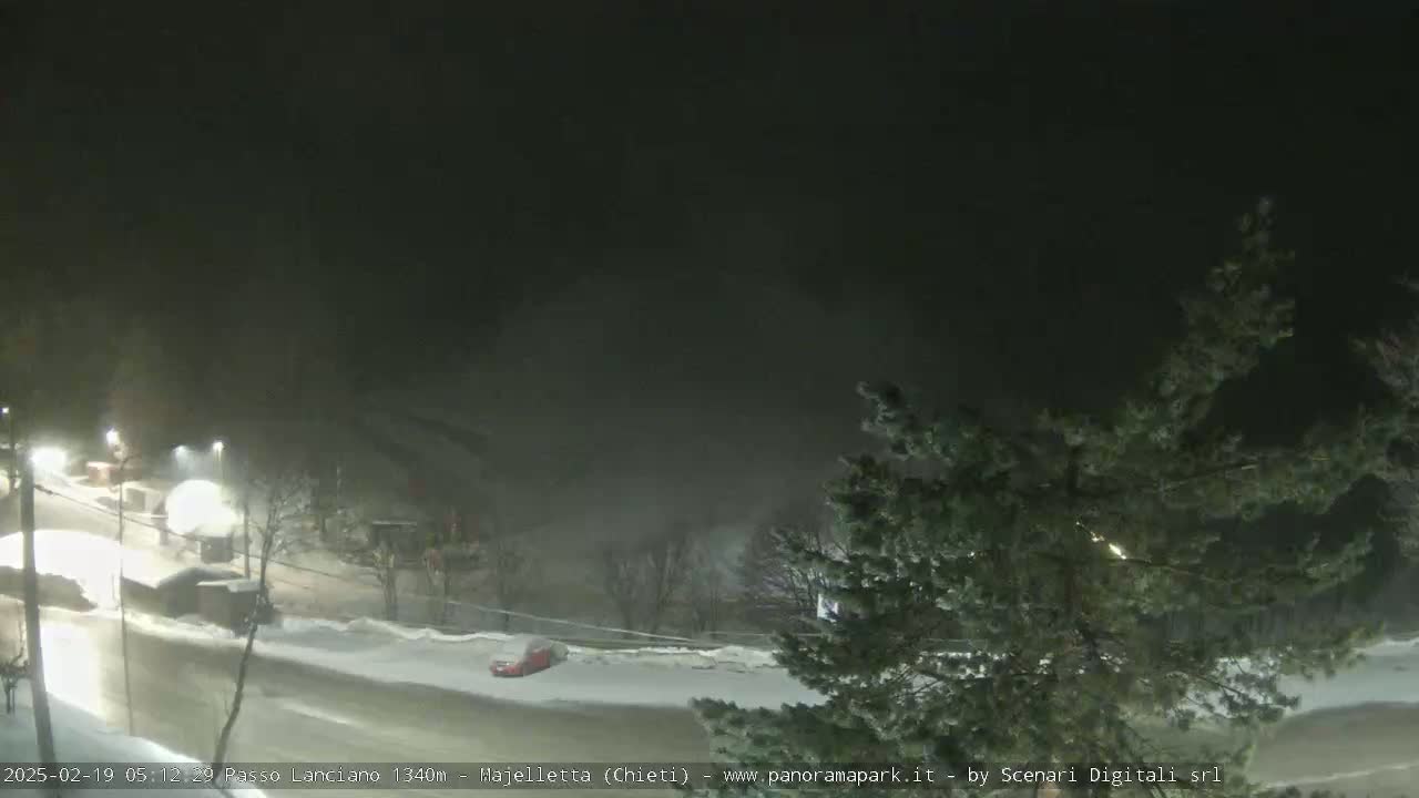 A nighttime, snowy scene shows a road with a few cars and buildings under a dark, foggy sky.