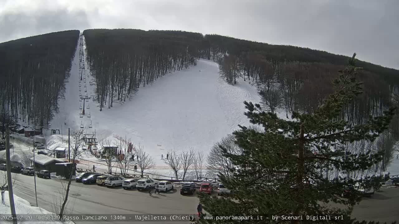 A ski slope with a chairlift and a parking lot of cars sits below a snow-covered hill covered in leafless trees under an overcast sky.