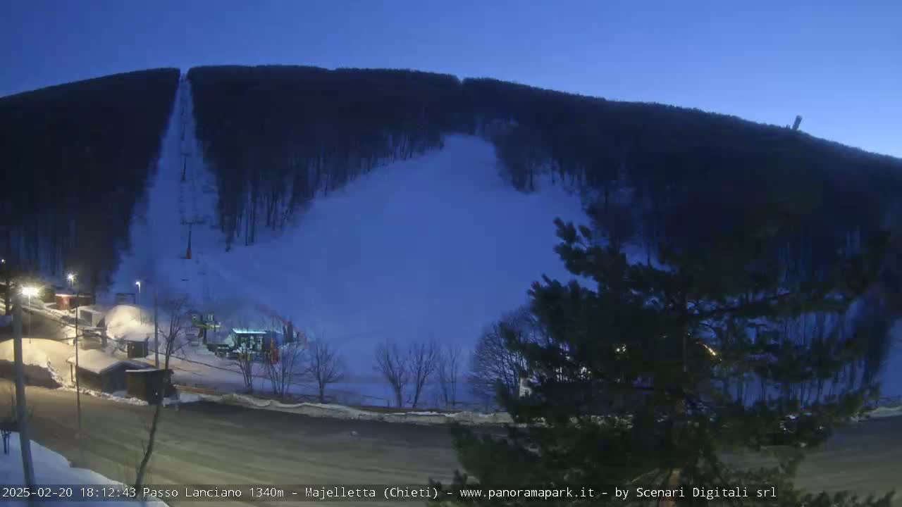 A snow-covered ski slope at twilight, with a chairlift visible, is flanked by dark, snow-dusted trees.