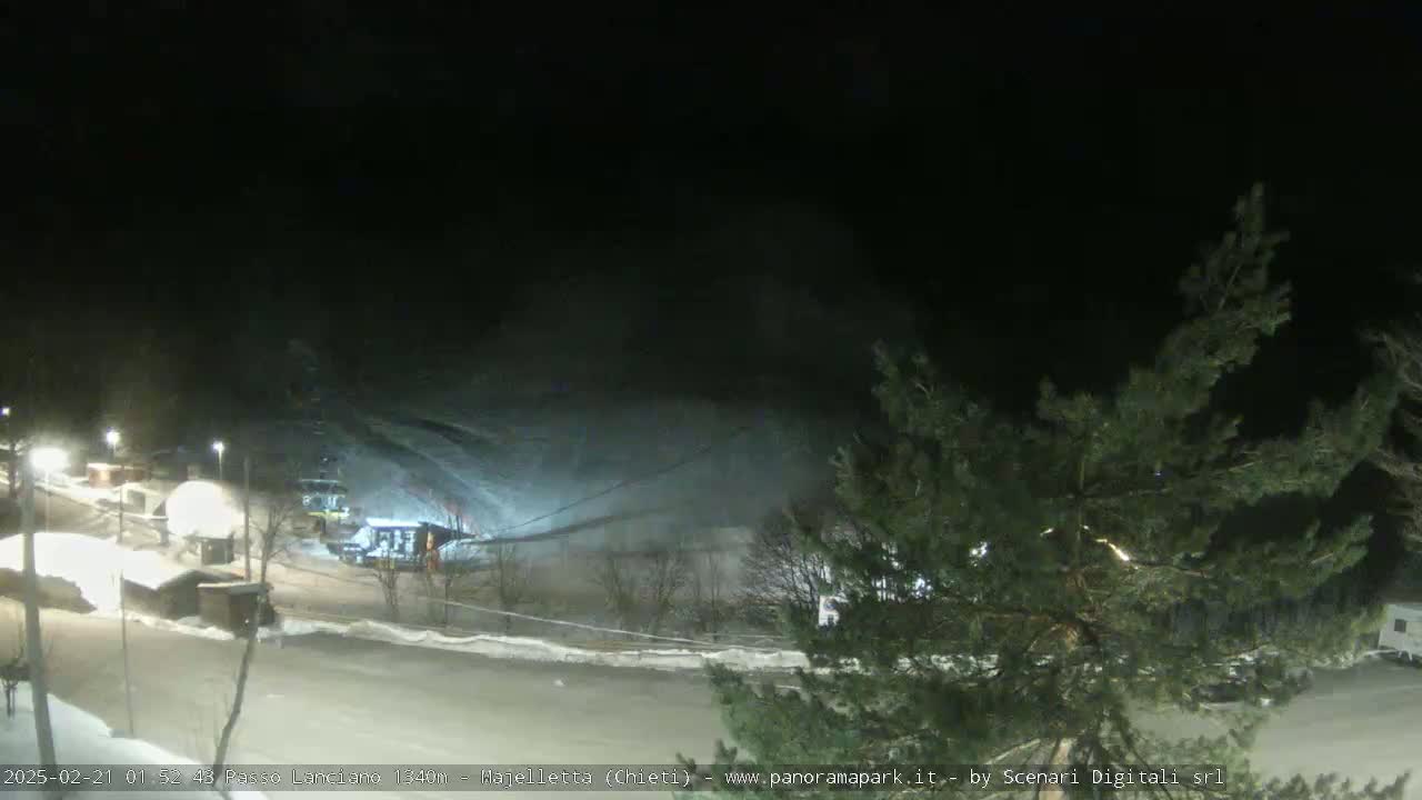A nighttime snow-covered scene shows a ski lift and several small buildings illuminated under a dark sky.