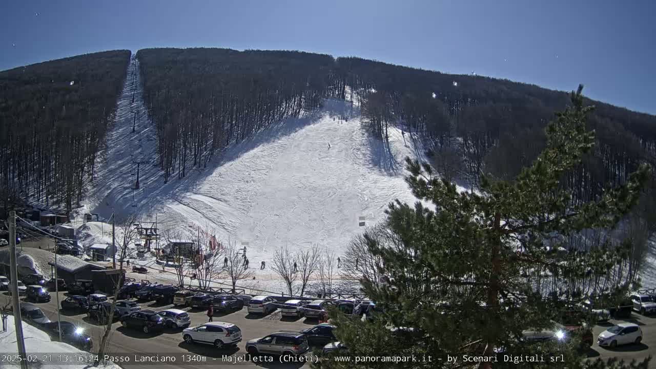 A snow-covered ski slope on a sunny day, with a parking lot full of cars at the base and a ski lift visible.
