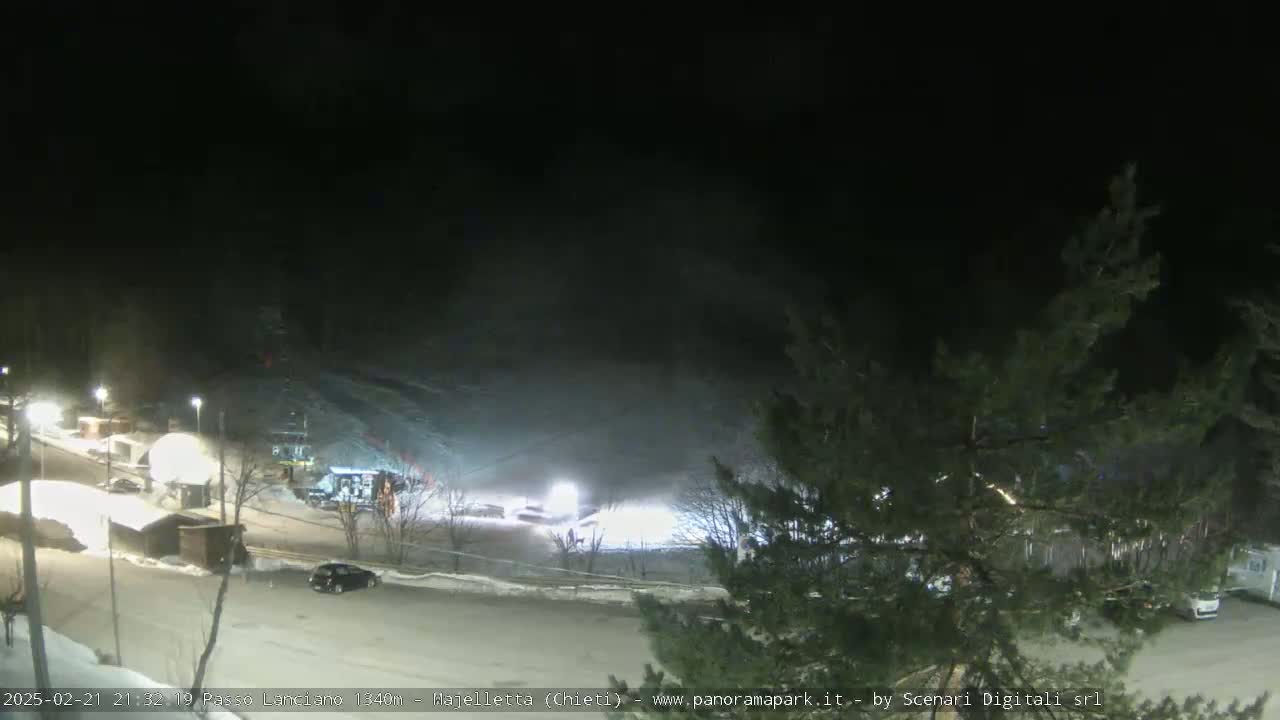 A nighttime, snow-covered parking lot and ski slope are illuminated, with a few vehicles visible.