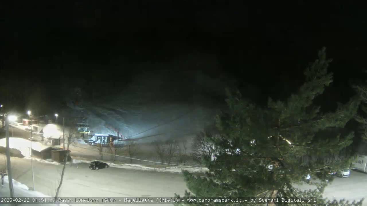 A nighttime, snowy scene shows a parking lot with vehicles near a ski lift and slope, under a dark sky.