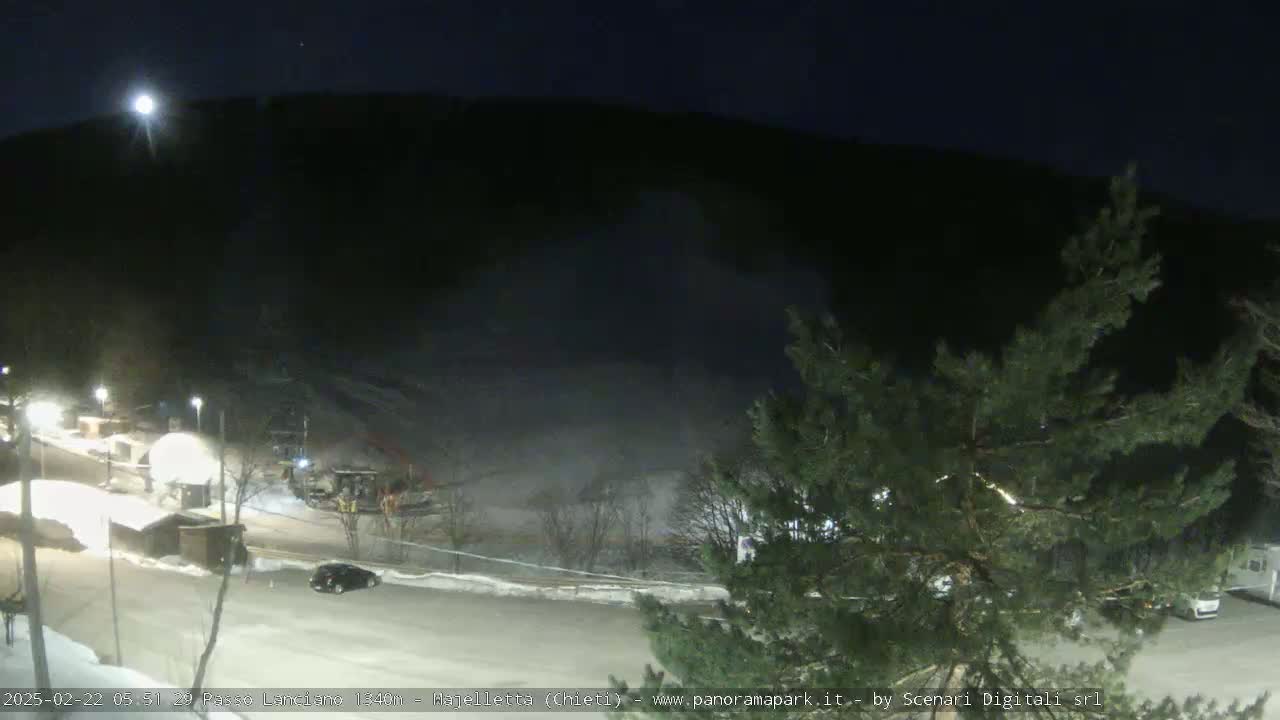 A nighttime view of a snow-covered parking lot and road, with a dark mountainside in the background under a moonlit sky.