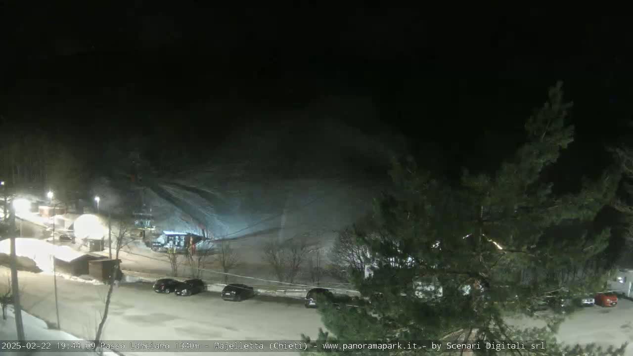 A nighttime view of a snow-covered ski slope with a parking lot and several cars visible below, under a dark sky.