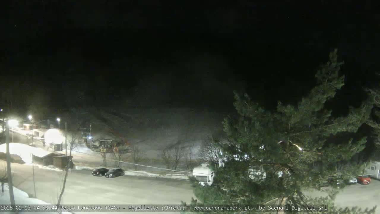 A nighttime view of a snow-covered parking lot with several vehicles, some small buildings, and trees, with snow faintly visible on the ground in the background.