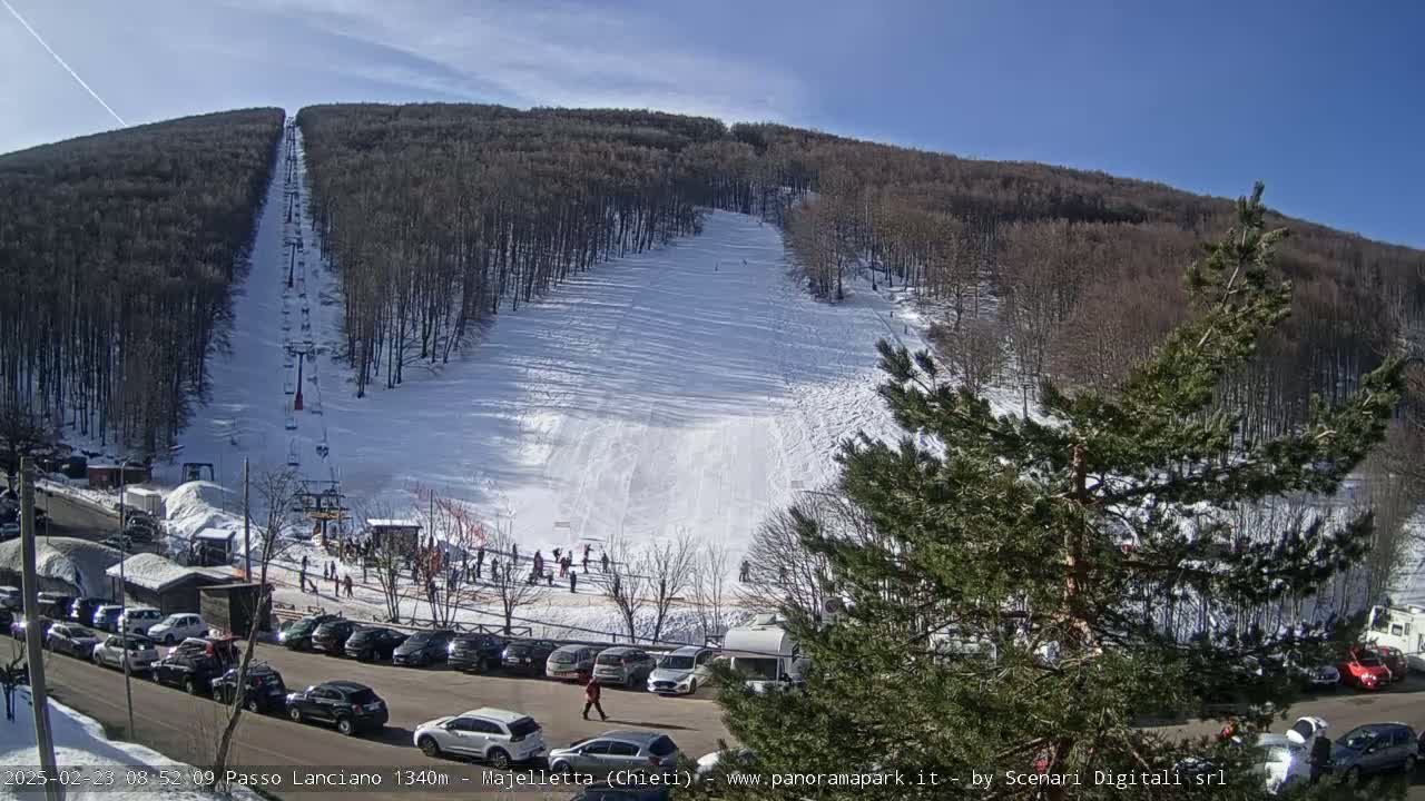 A ski slope on a sunny day with a chairlift visible, surrounded by snow-covered trees and a parking lot full of cars.