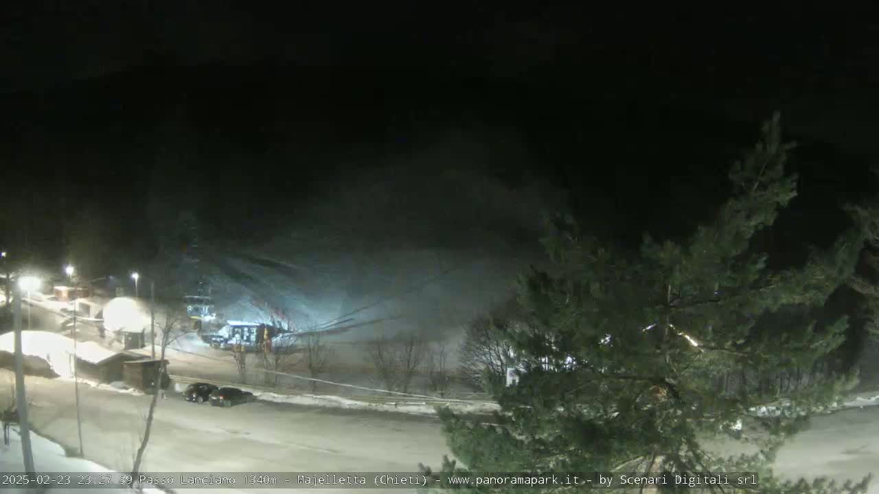 A nighttime, snow-covered ski slope with several cars parked in the lower area, under a dark, snowy sky.