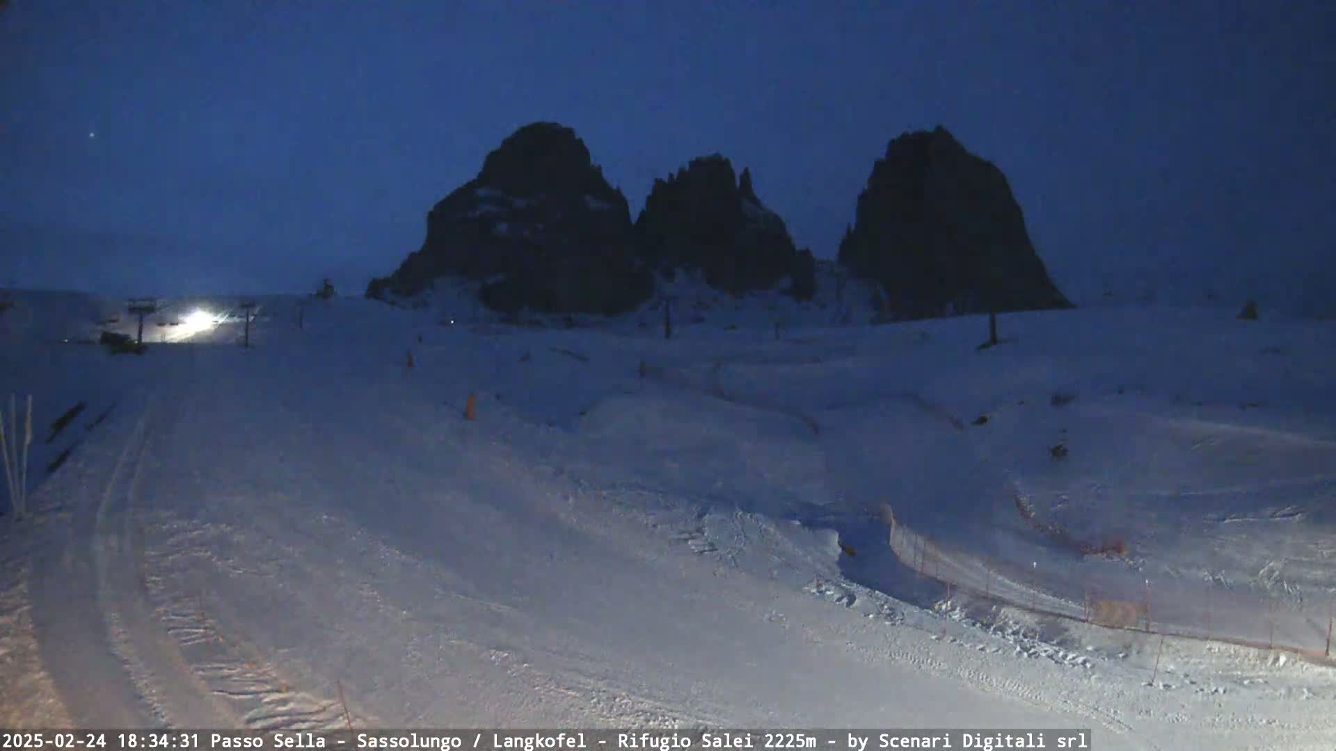 A snow-covered landscape at twilight, featuring three dark, rocky peaks in the background and a ski slope in the foreground with some ski lifts.