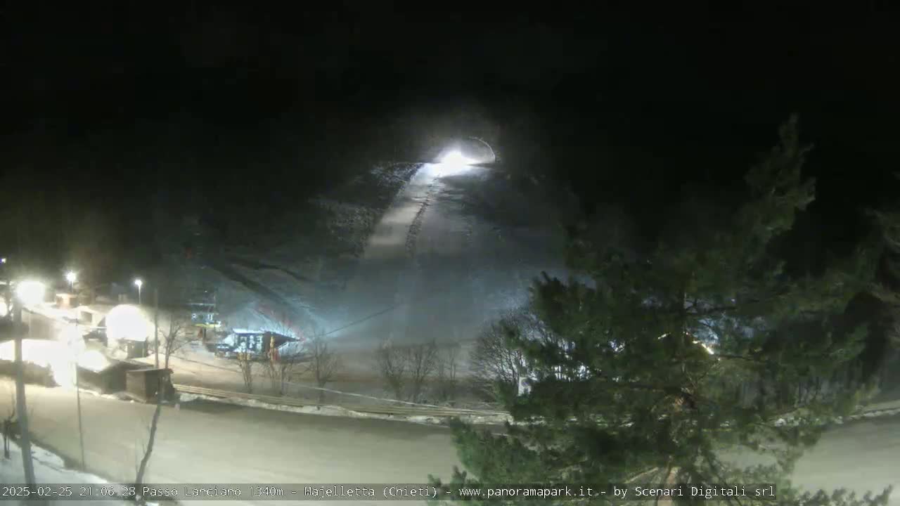 A snow-covered ski slope is illuminated at night, with a tunnel-like structure at the top and some buildings visible at the bottom.