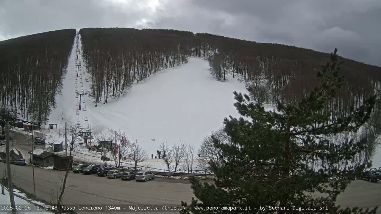A snow-covered ski slope with a chairlift and several cars parked in a lot under an overcast sky.
