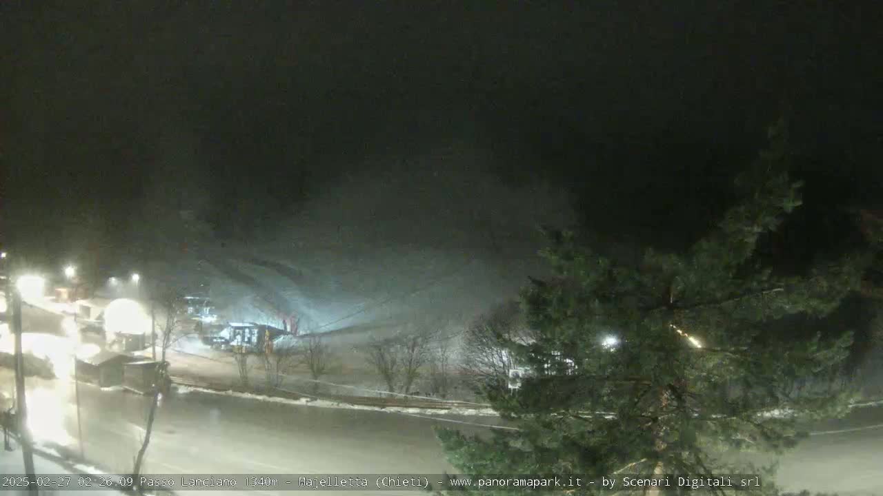 A nighttime view of a snow-covered landscape, with falling snow and illuminated buildings and roads.