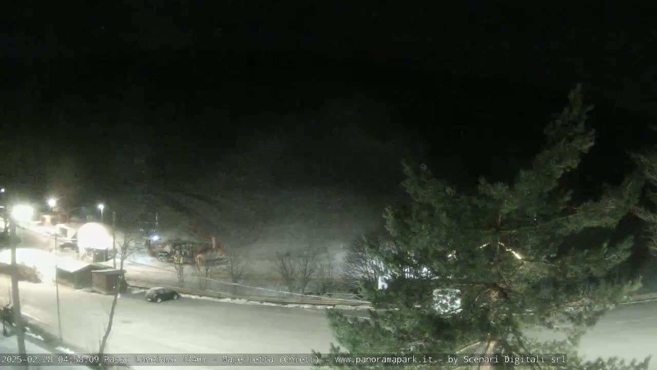 A nighttime, snow-covered parking lot with a few vehicles and buildings visible under a dark sky.