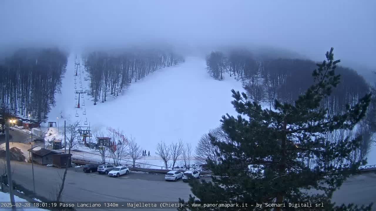 A foggy, snowy ski slope with a chairlift and a parking lot below, surrounded by snow-covered trees.