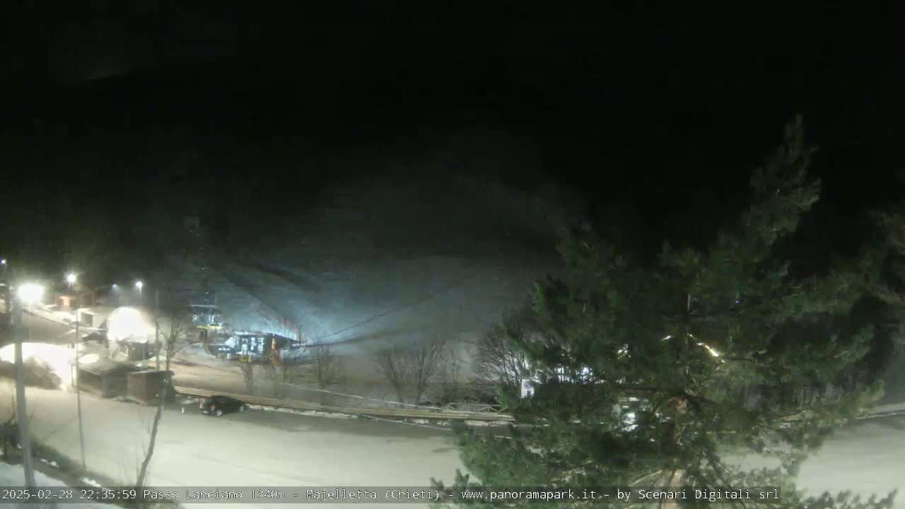A nighttime, snow-covered ski resort scene with a few vehicles parked near buildings and some lights illuminating the area.