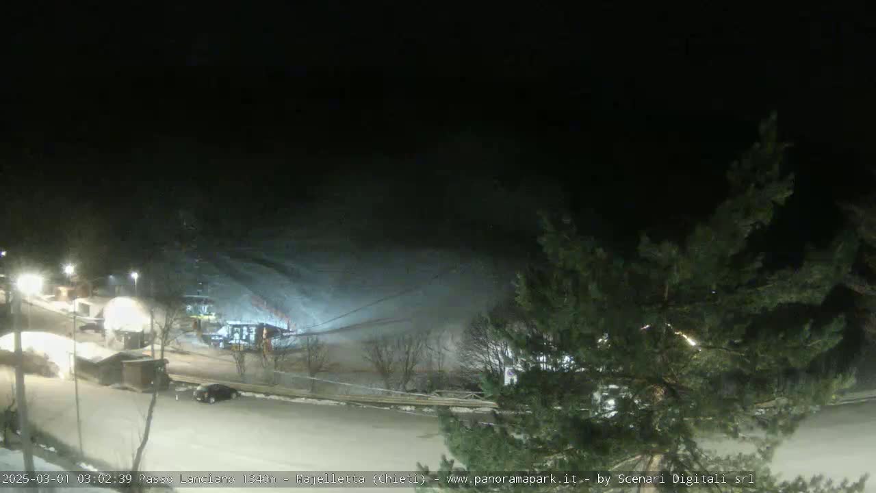 A nighttime view of a snow-covered ski slope with a few cars and small buildings visible in the foreground, under dark skies.