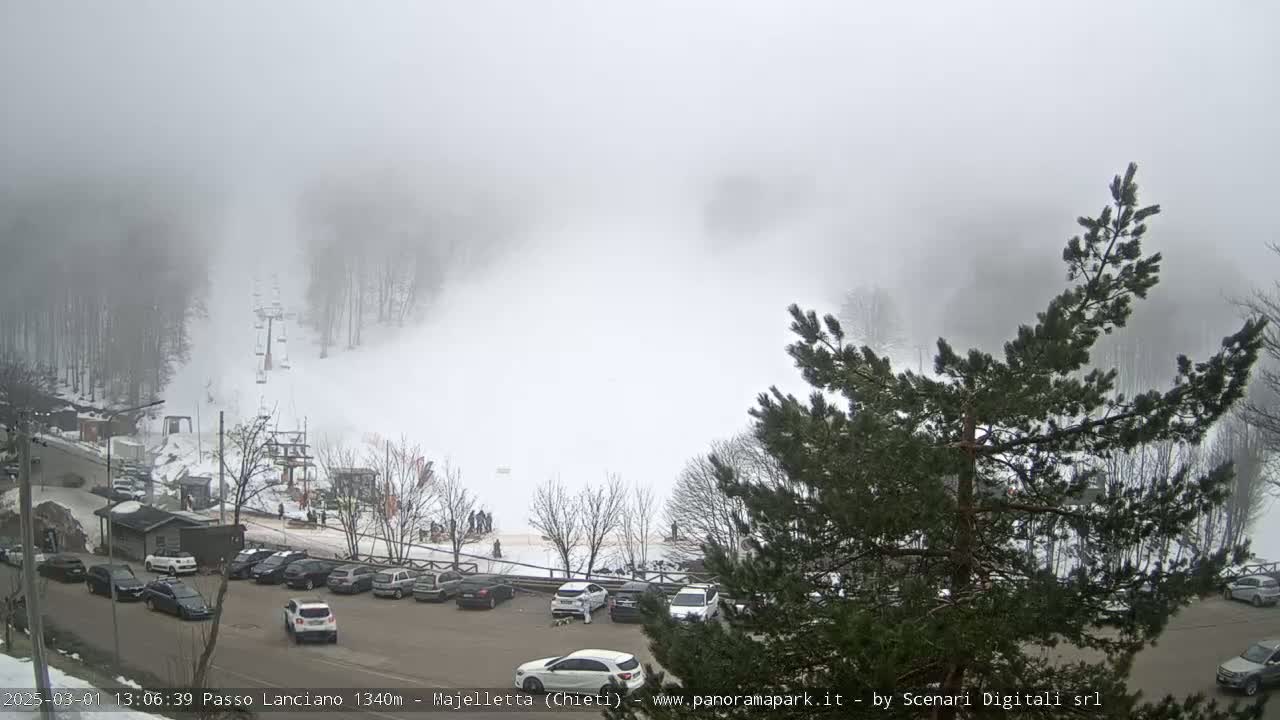 A snowy ski slope is shrouded in thick fog, with a parking lot full of cars visible in the foreground.