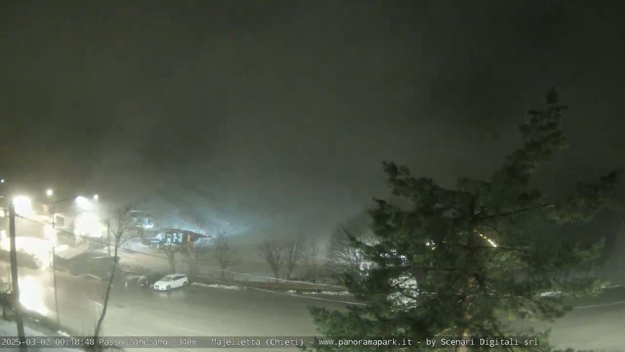 A nighttime scene of a snow-covered parking lot and road, shrouded in heavy fog.