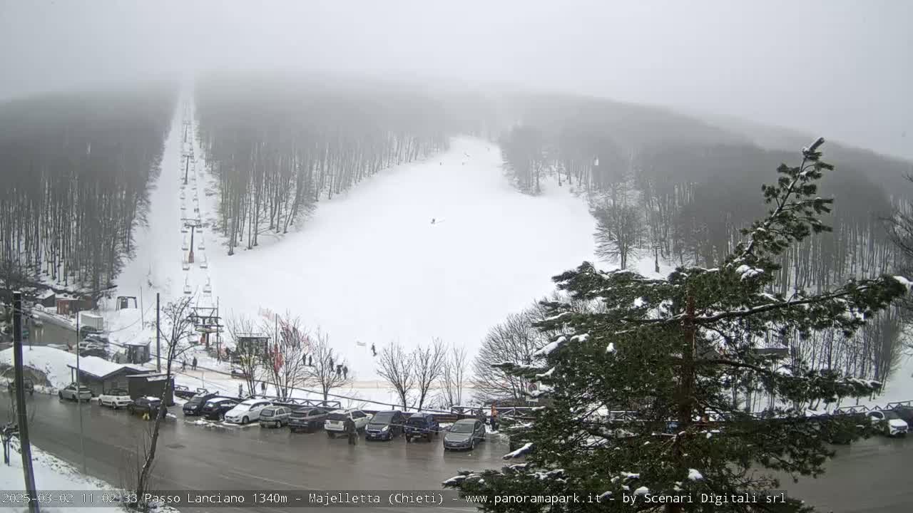 A snowy ski slope with a chairlift and a parking lot is visible under foggy conditions.