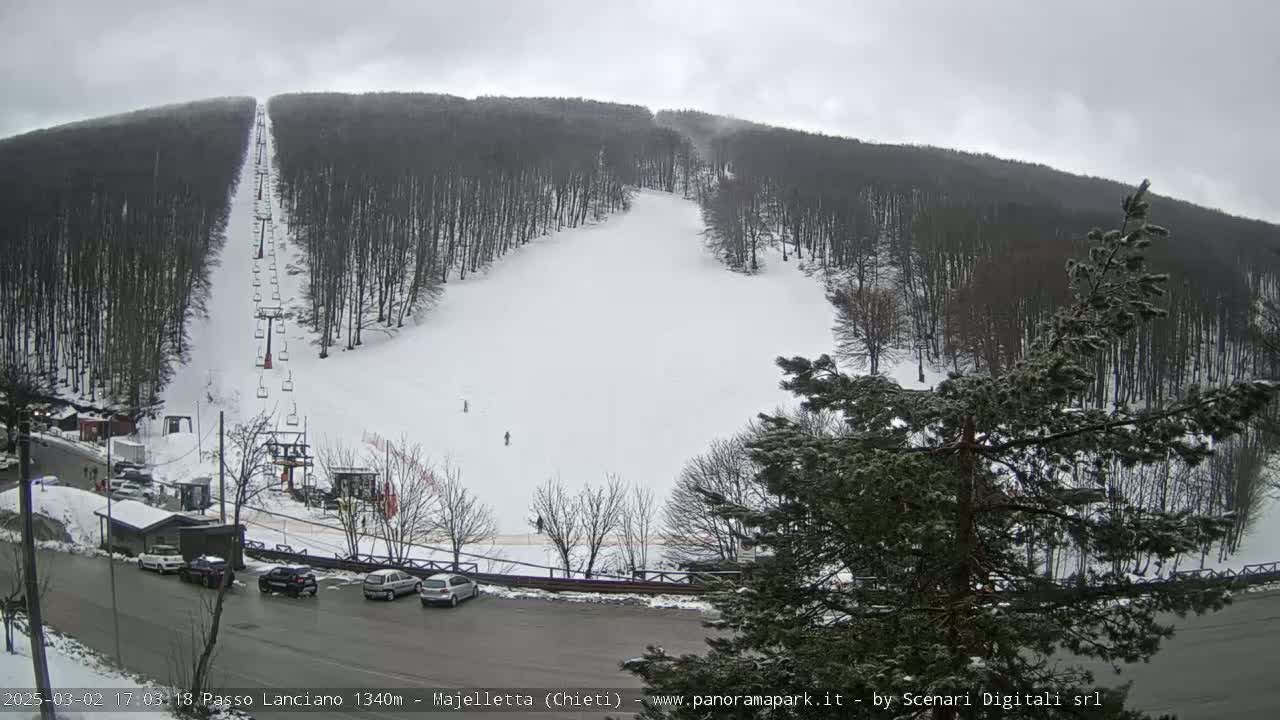 A snow-covered ski slope with a chairlift and a few skiers is visible under an overcast sky, with cars parked in a lot at the base of the hill.