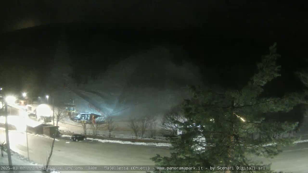 A nighttime view of a snow-covered ski slope with several buildings and vehicles visible under falling snow.