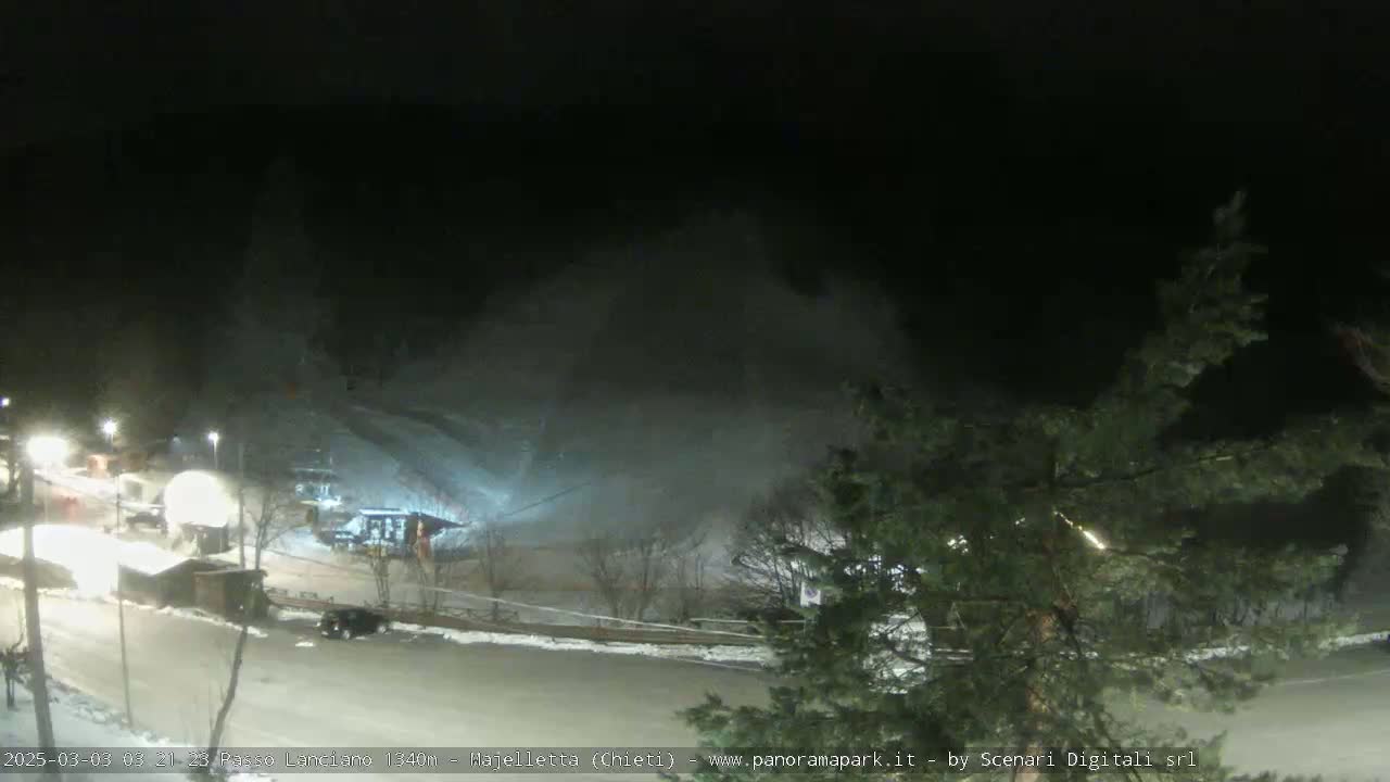 A snowmaking machine is operating at night on a ski slope, illuminating the area with bright white snow.