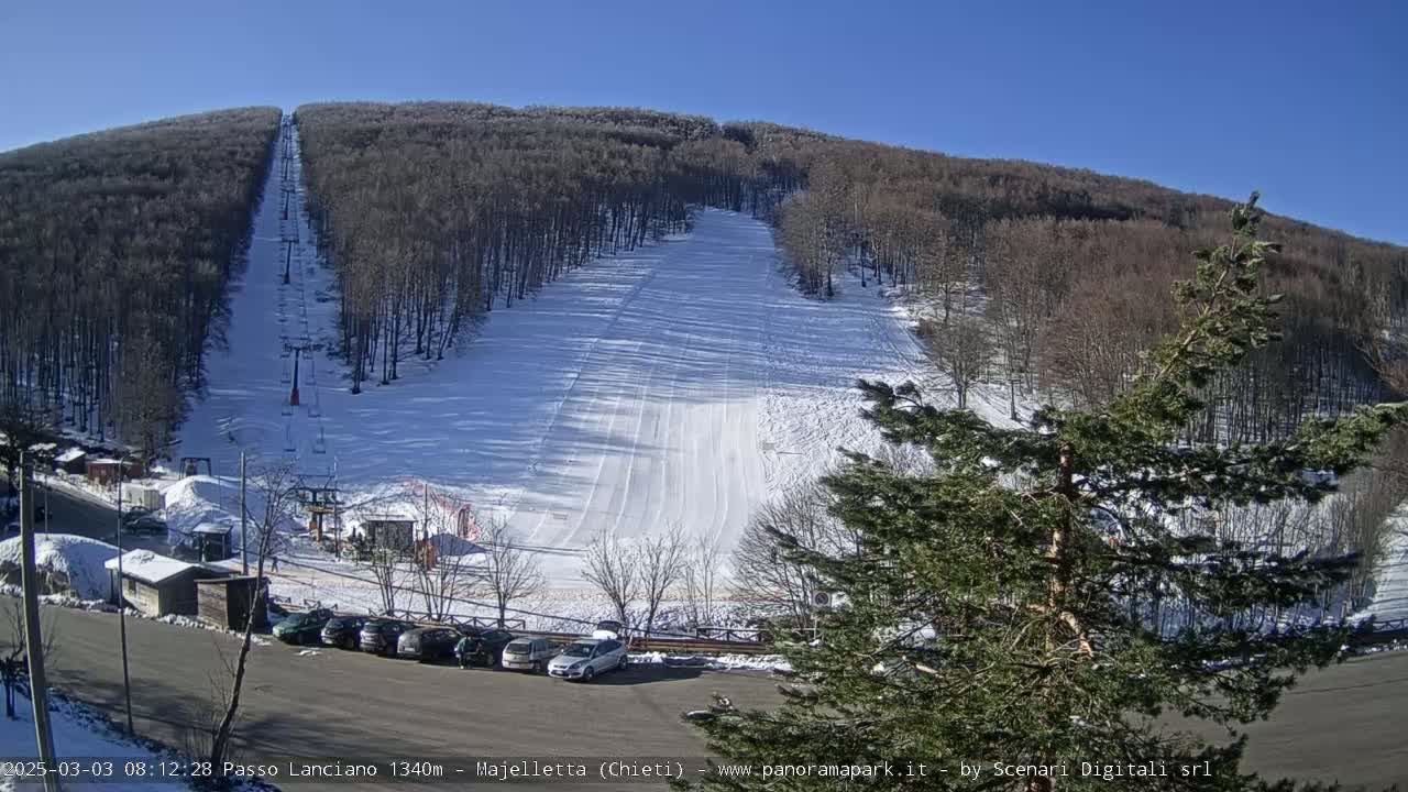 A ski slope on a snow-covered hillside under a clear blue sky, with a chairlift and a parking lot with several cars visible at the bottom.