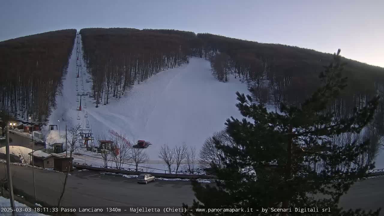 A snow-covered ski slope with a chairlift and a snow groomer is visible under a twilight sky.