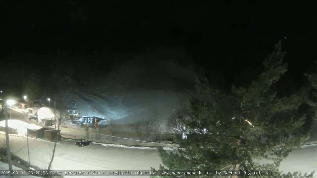 A nighttime view of a snow-covered parking lot and ski slope, illuminated by artificial lights under a dark sky.