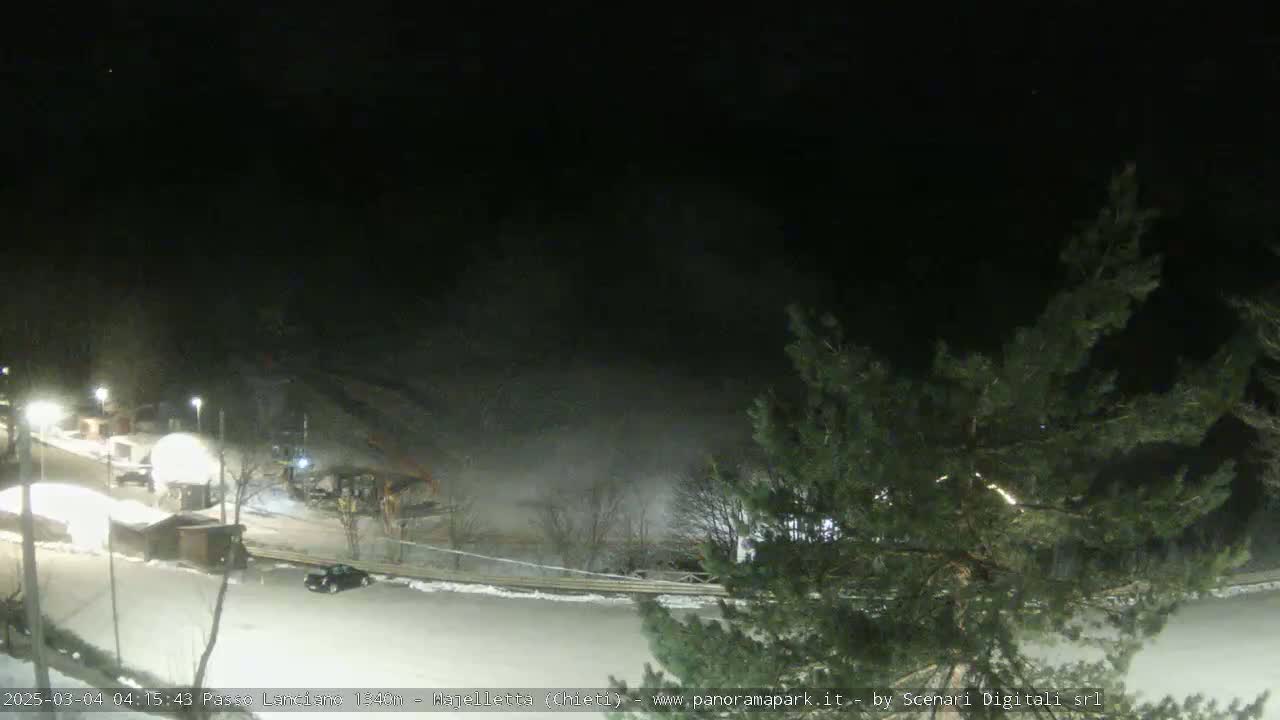 A nighttime, snow-covered parking lot and surrounding area with some lights and a few vehicles, partially obscured by fog or mist.