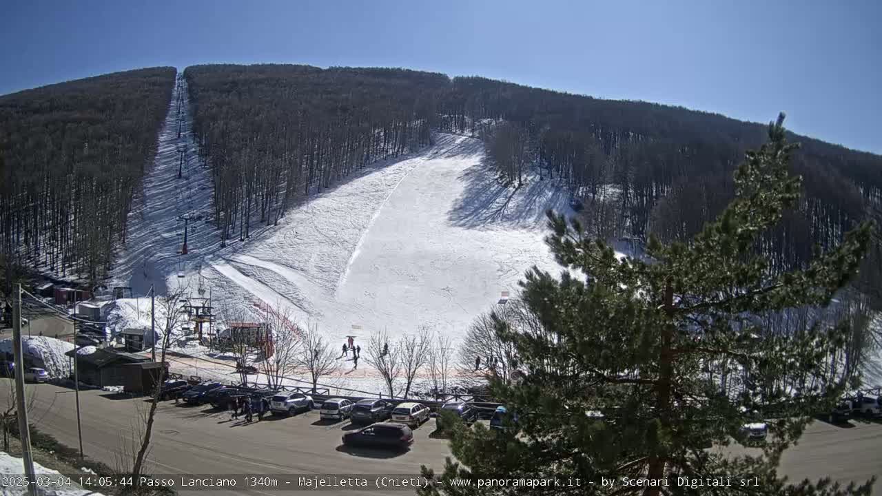 A ski slope with a chairlift and a parking lot full of cars is visible under a sunny sky.