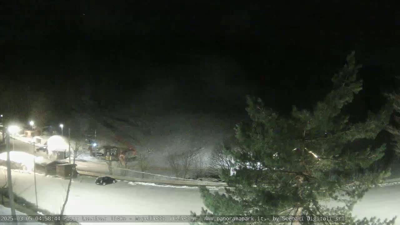 A nighttime, snow-covered parking lot and roadway are partially visible beneath dark, leafless trees, with several cars and small structures illuminated by artificial lights.