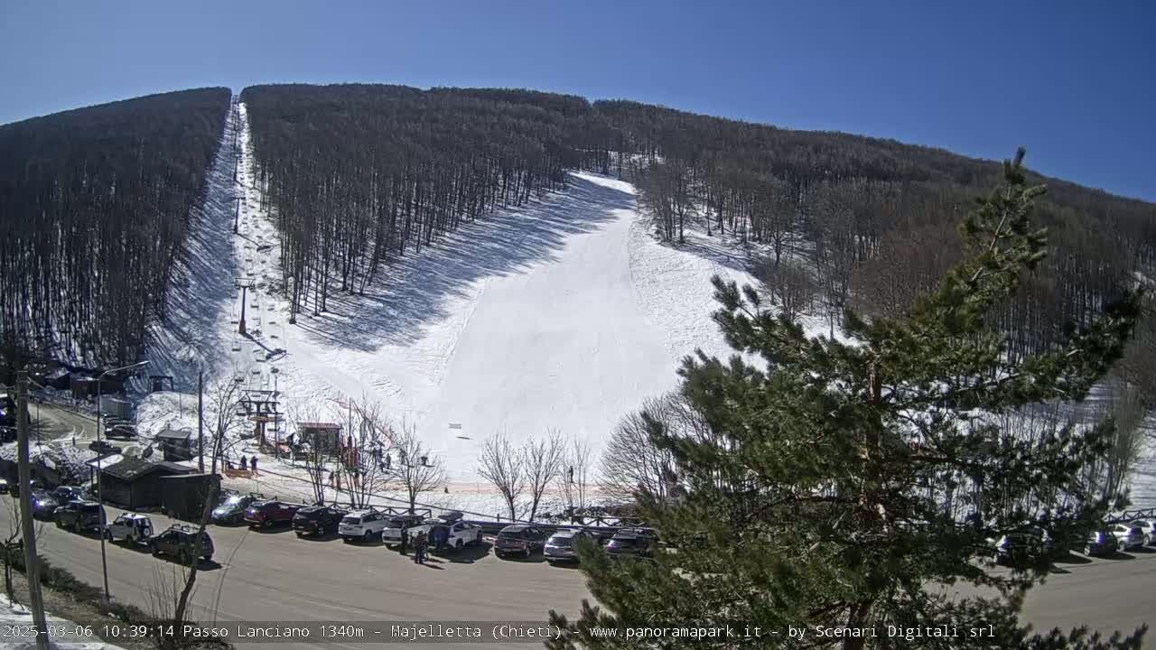 A ski slope with a chairlift and a parking lot full of cars is visible under a sunny sky.