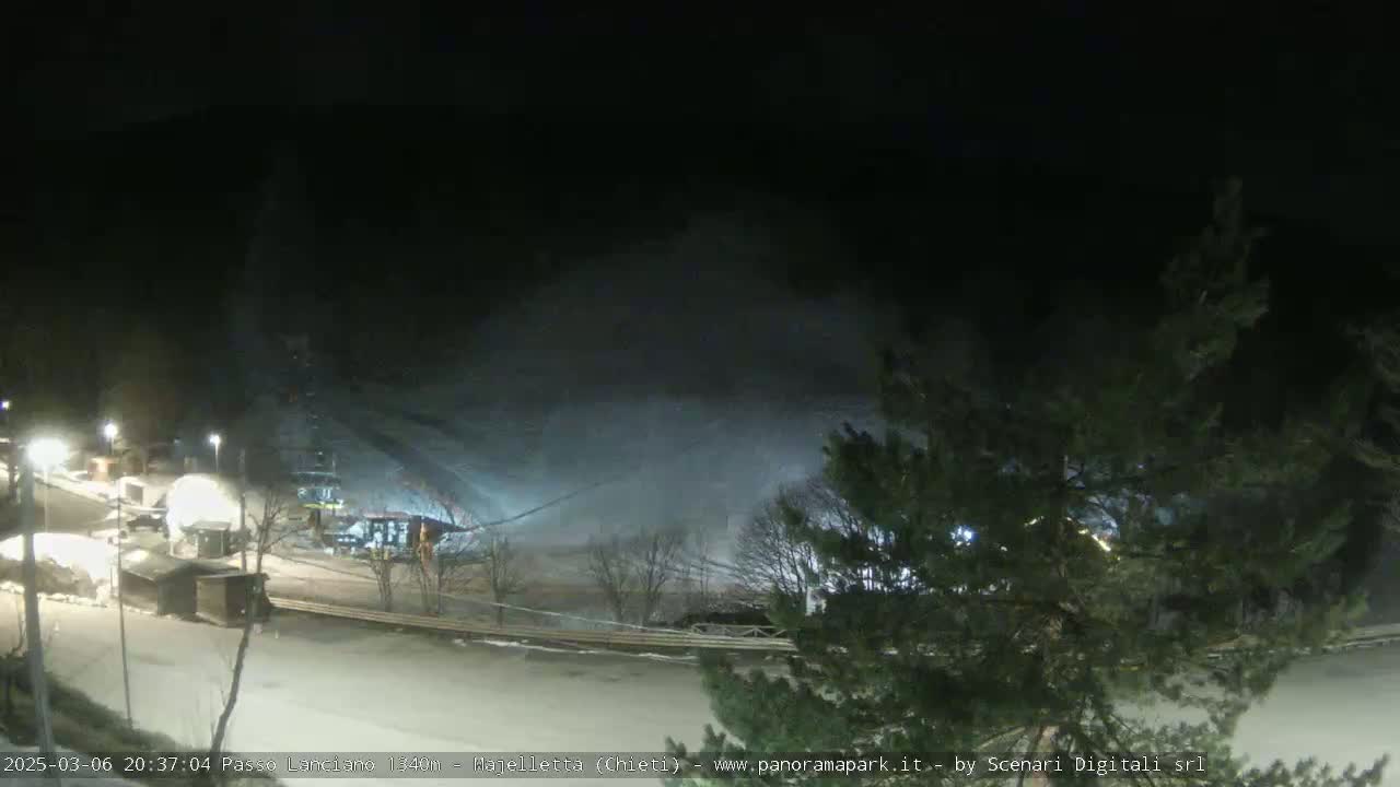 A nighttime view of a snow-covered ski slope with snow blowing across it, illuminated by artificial lights.