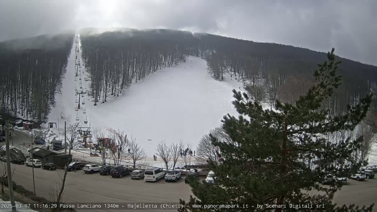 A snow-covered ski slope with a chairlift and several skiers, situated within a wooded mountain area under an overcast sky.