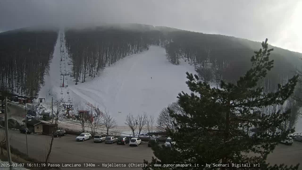 A snowy ski slope with a chairlift and several cars parked at the bottom, under an overcast sky.