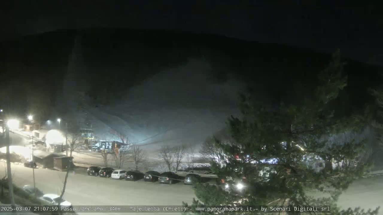A nighttime snow scene shows a snow-covered hillside with a parking lot containing several cars below, illuminated by artificial lights.