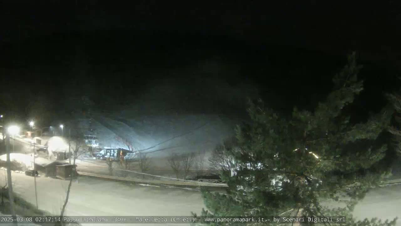 A snow-covered ski slope at night, illuminated by artificial lights, with some small buildings and trees visible.