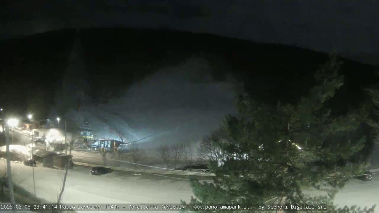 A nighttime view of a snow-covered ski area with a lift and some buildings, under heavy snowfall.