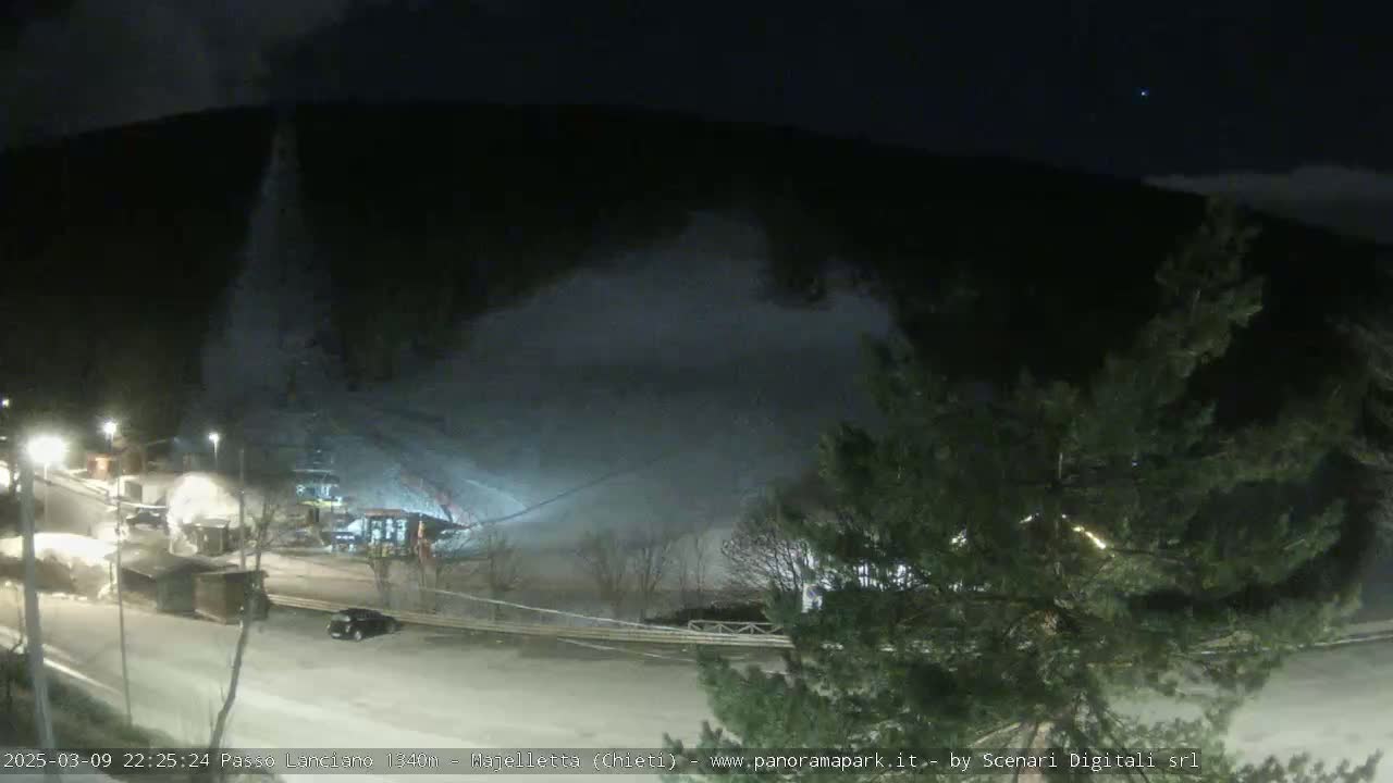 A nighttime view of a snow-covered area, possibly a ski resort, with artificial lights illuminating a road and some buildings under a partly cloudy sky.