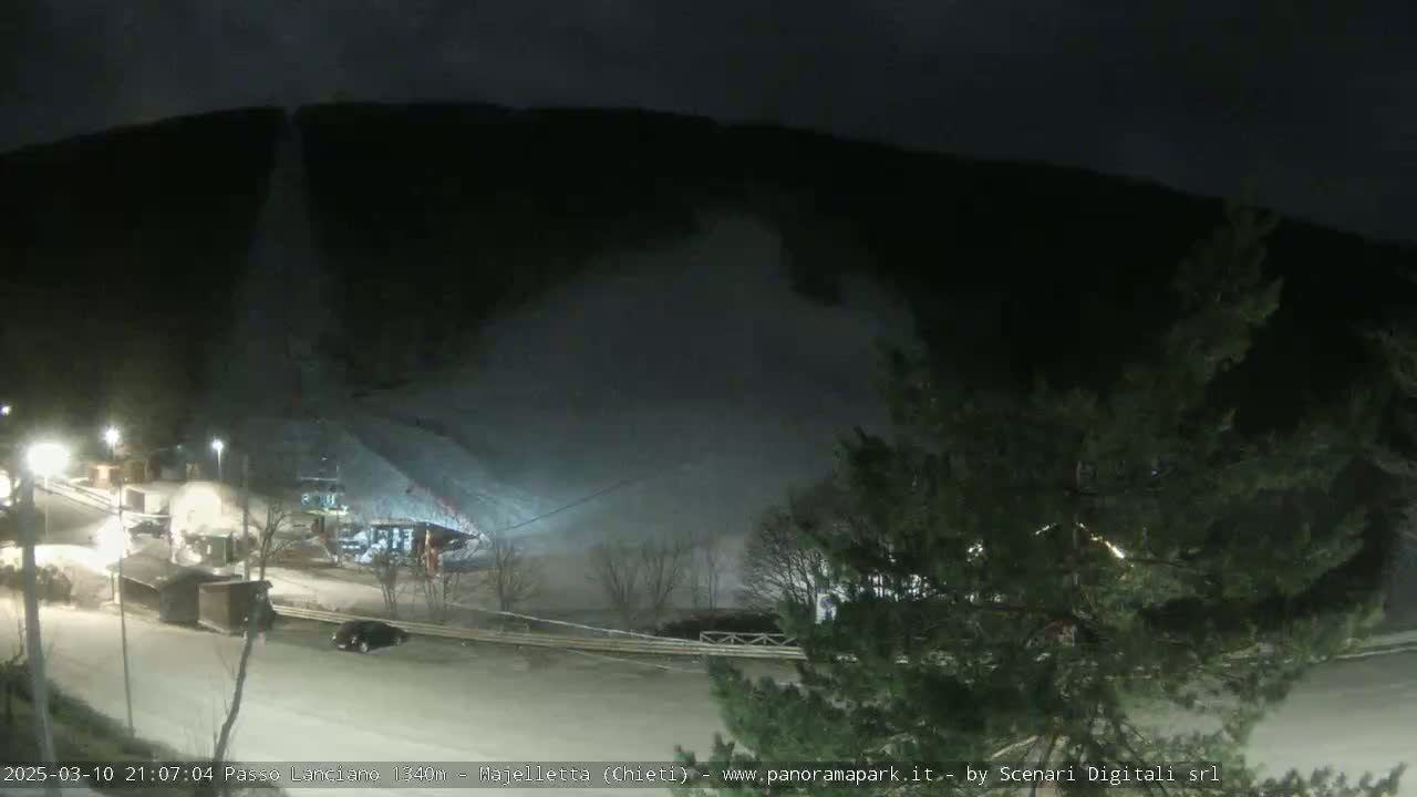 A nighttime scene shows a snow-covered hillside with a plume of snow blowing across a road at the base of the hill, and several vehicles and structures are visible below.
