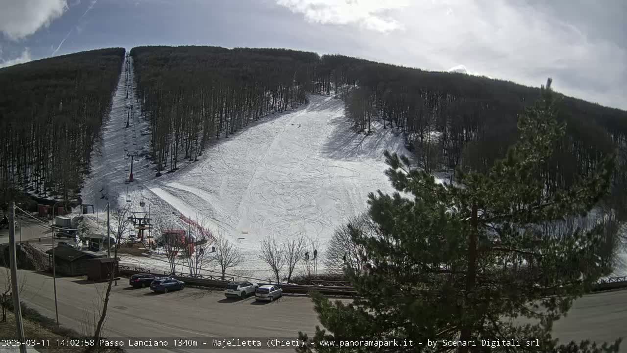 A snowy ski slope with a chairlift is visible on a sunny day, surrounded by a dark forest and a small parking lot with several cars.