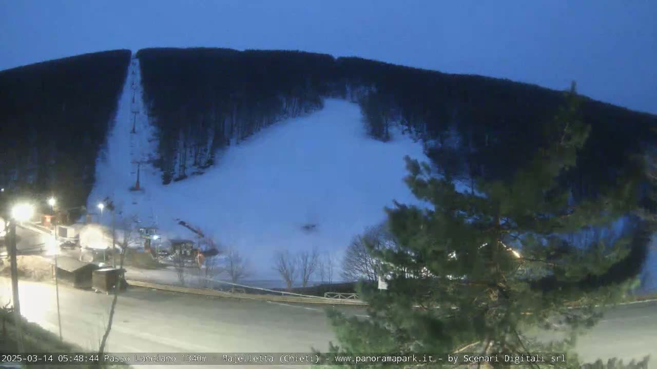 A snow-covered ski slope at twilight, illuminated by artificial lights, is flanked by dark trees and a road.