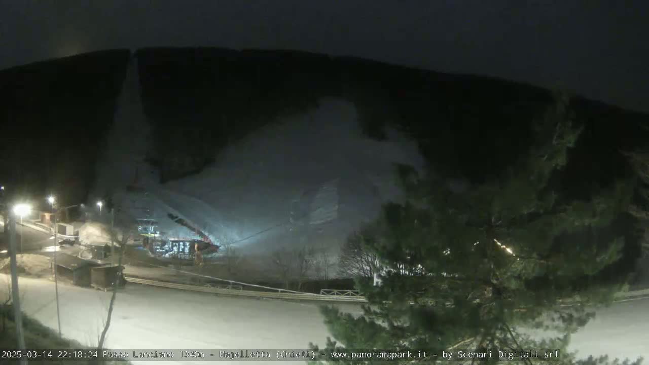 A nighttime, snow-covered hillside is illuminated by artificial lights, with a road and some structures visible in the foreground.
