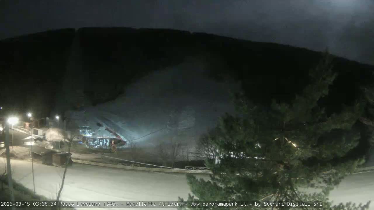 A nighttime view of a snow-covered slope and road, with some buildings and trees visible under a dark sky.