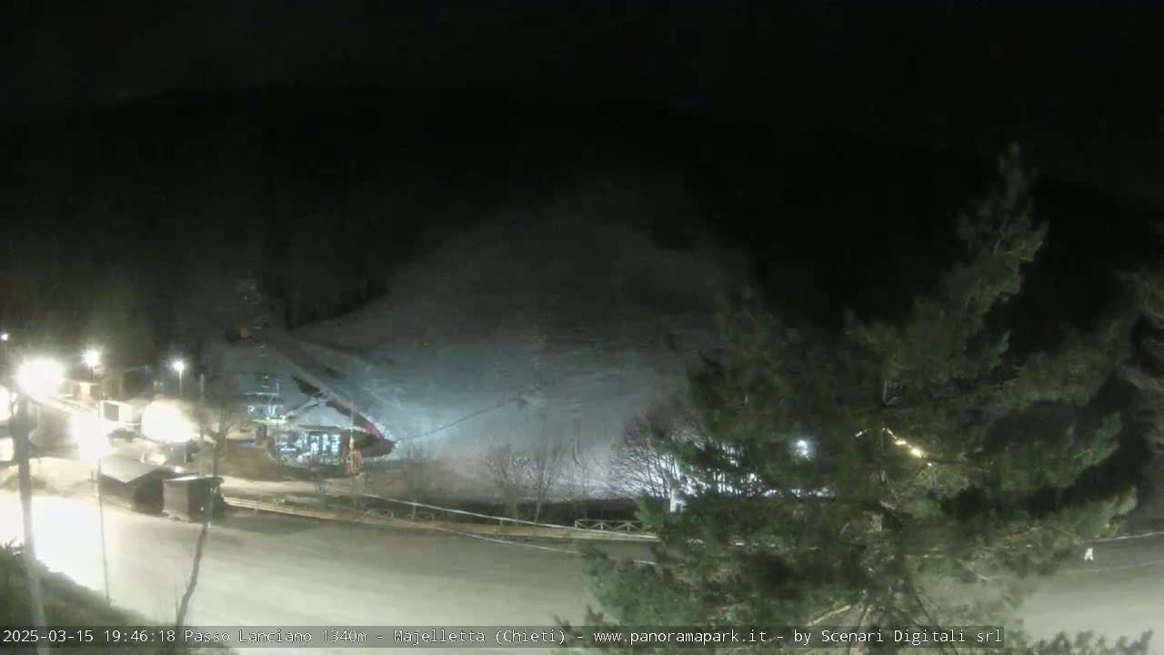A nighttime, snow-covered slope is illuminated by artificial lights, with some buildings and trees visible in the foreground.
